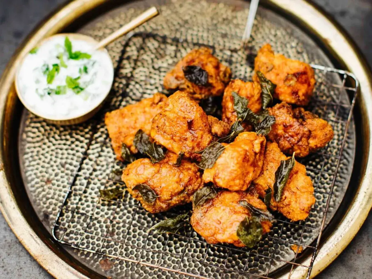 Knusprige chicken pakora, garniert mit frischen Kräutern, auf einem Gitterrost serviert. Daneben eine Schale mit Joghurt-Dip.