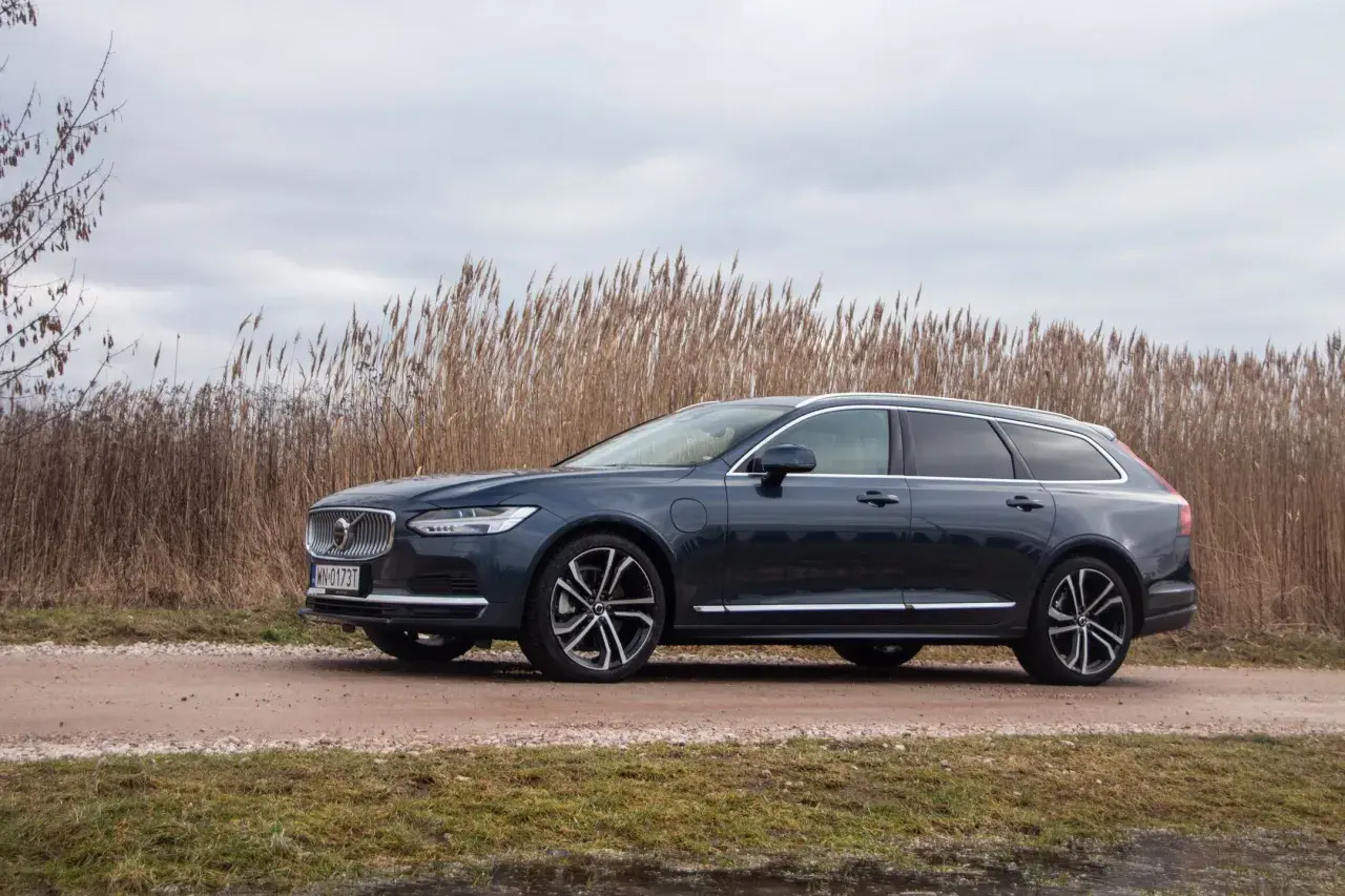 Ciemnogranatowe Volvo v90 na tle wysokiej, suchej trawy i pochmurnego nieba.