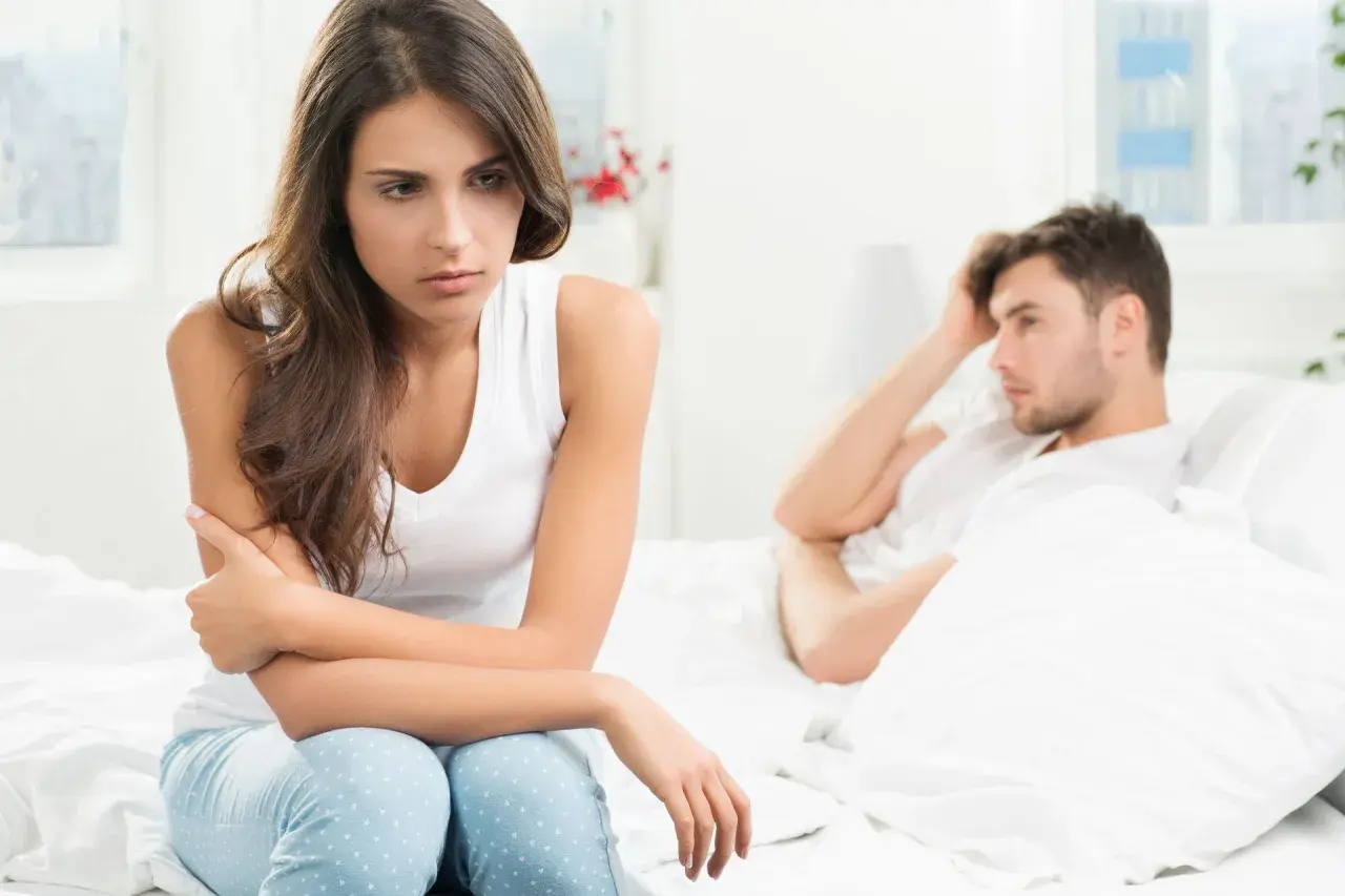 Smutna kobieta siedzi na łóżku, zmartwiona. Jej partner leży obok, z ręką na czole. Czy to uczulenie na spermę powoduje ich problemy?