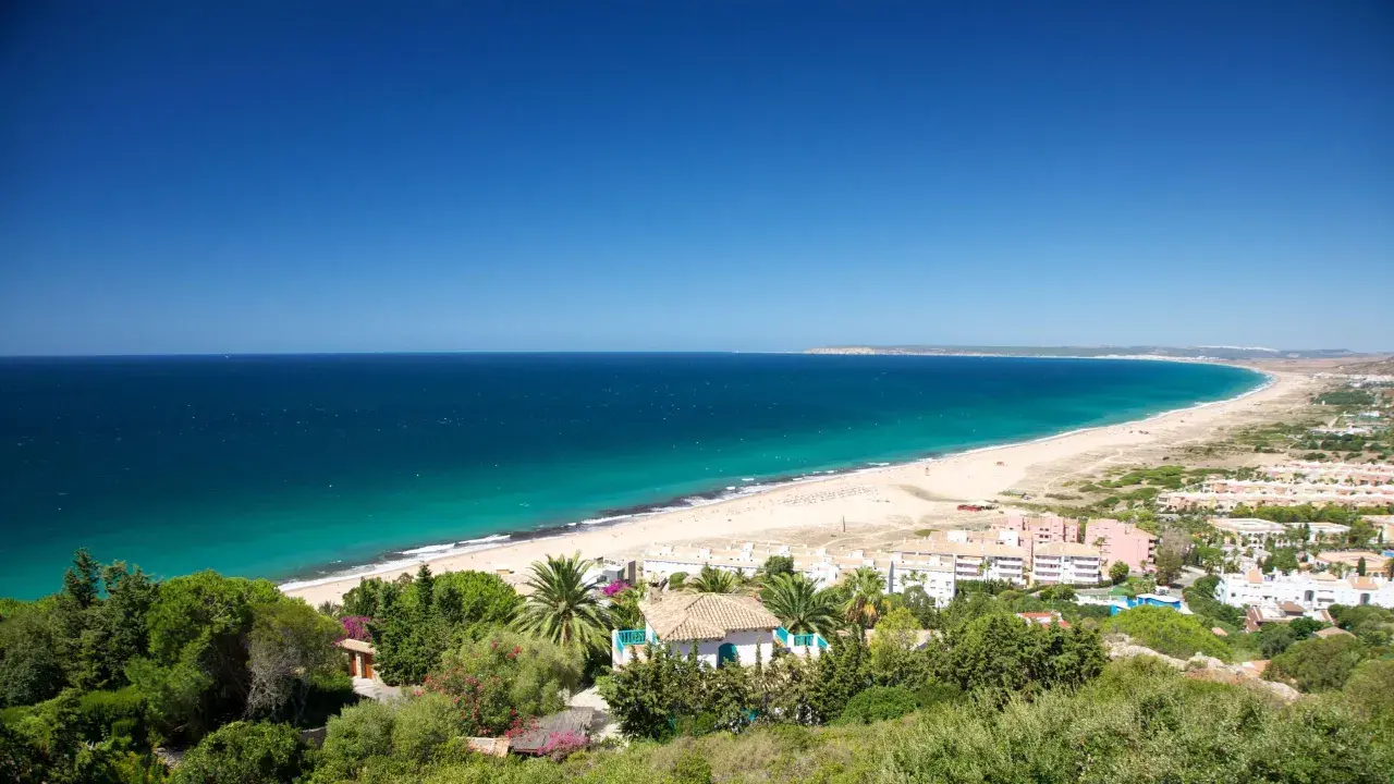 Vista panorámica del pueblo y paseo marítimo de Zahara de los Atunes, con su extensa playa de arena dorada y el mar azul turquesa.