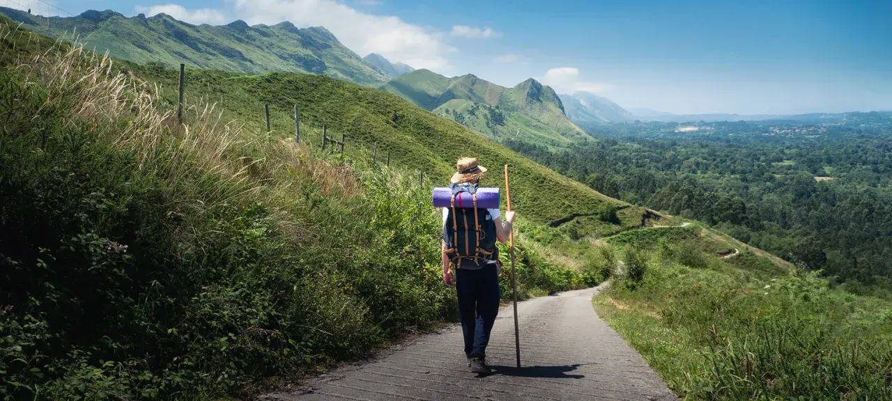 Pielgrzym w kapeluszu i z plecakiem idzie ścieżką przez zielone wzgórza. To jedna z malowniczych tras Camino de Santiago.
