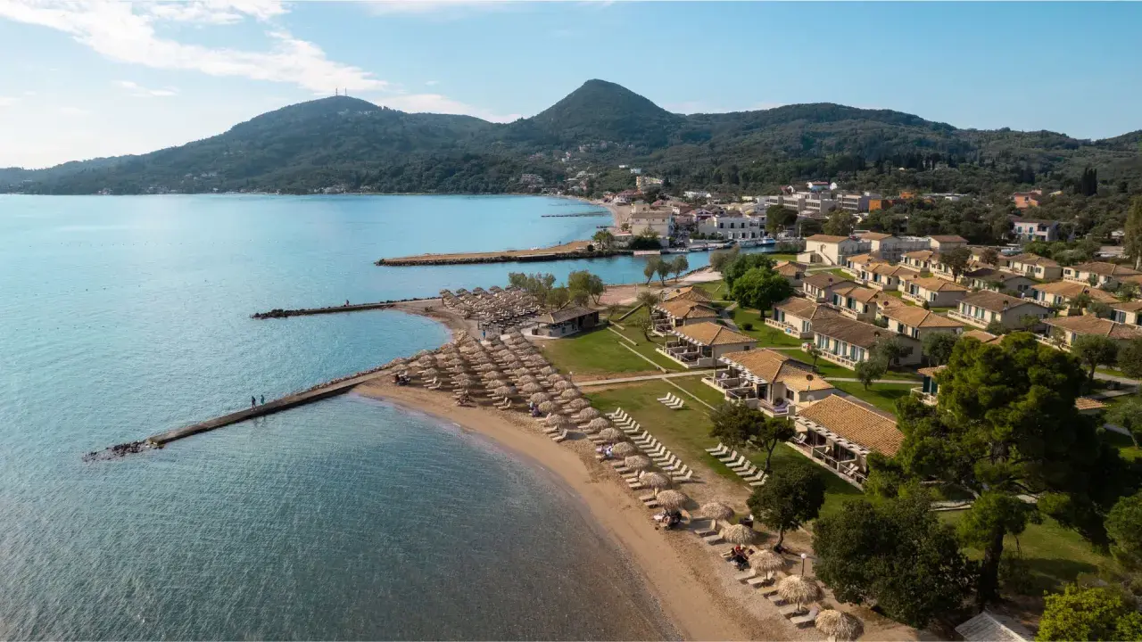 Piękna plaża z rzędami leżaków i parasoli, domki z dachówką, góry w tle. Idealne miejsce na wakacje, jak sugerują messonghi beach holiday resort opinie.