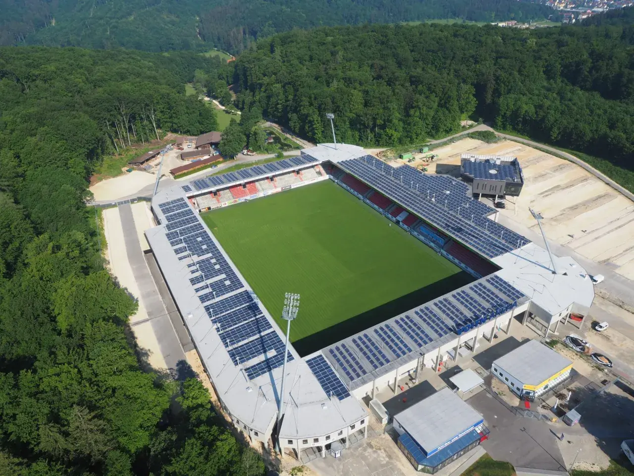 Widok z lotu ptaka na zieloną murawę i trybuny stadionu, otoczonego lasem. To najmniejszy stadion na świecie, z panelami słonecznymi na dachu.