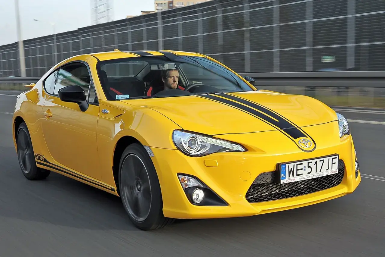 Żółte coupé Toyota GT86 z czarnymi pasami na masce jedzie po drodze.