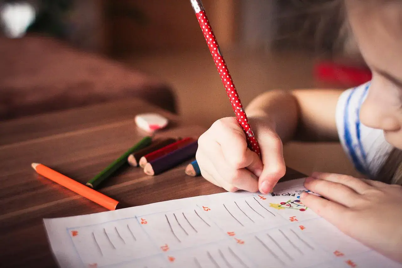 Bambina scrive con matita a pois rossi, circondata da pastelli. Forse sta calcolando l'indennità di frequenza scolastica importo.