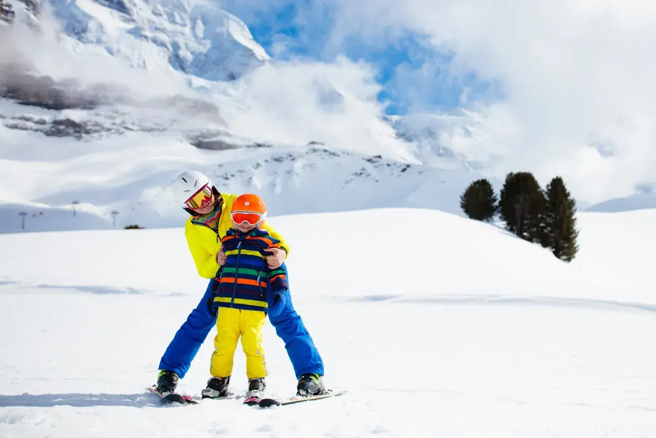 Un adulto insegna a sciare a un bambino di 2 anni su una pista innevata, con montagne sullo sfondo. Un perfetto weekend con bambini 2 anni!