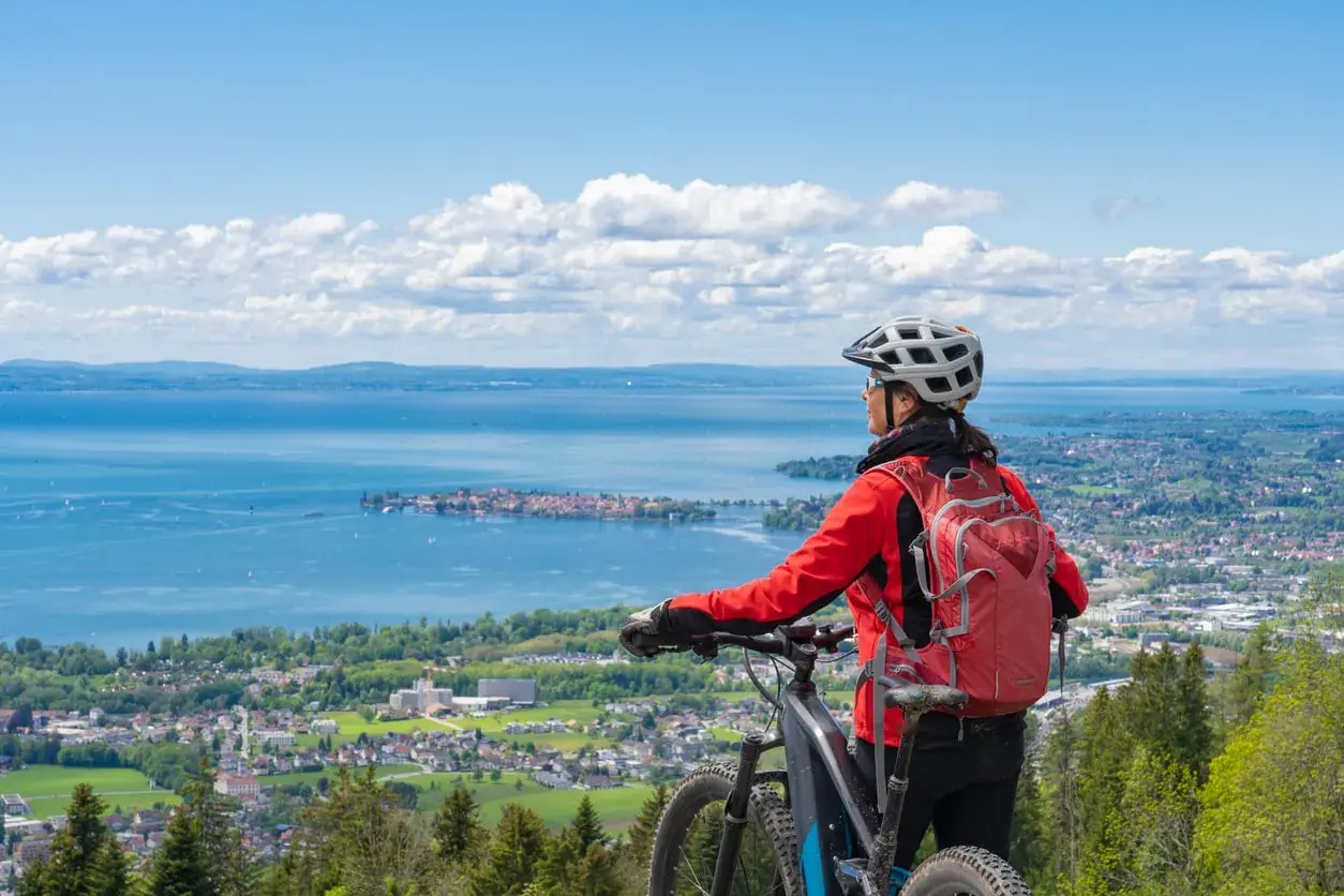 A woman in a red jacket and helmet pauses her **Lake Constance bike tour** to admire the expansive blue lake and distant towns under a cloudy sky.
