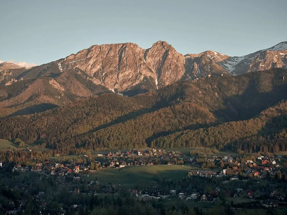 Widok z **Antałówki punkt widokowy** na Tatry. Słońce oświetla skaliste szczyty i lasy, a w dole widać zabudowania.