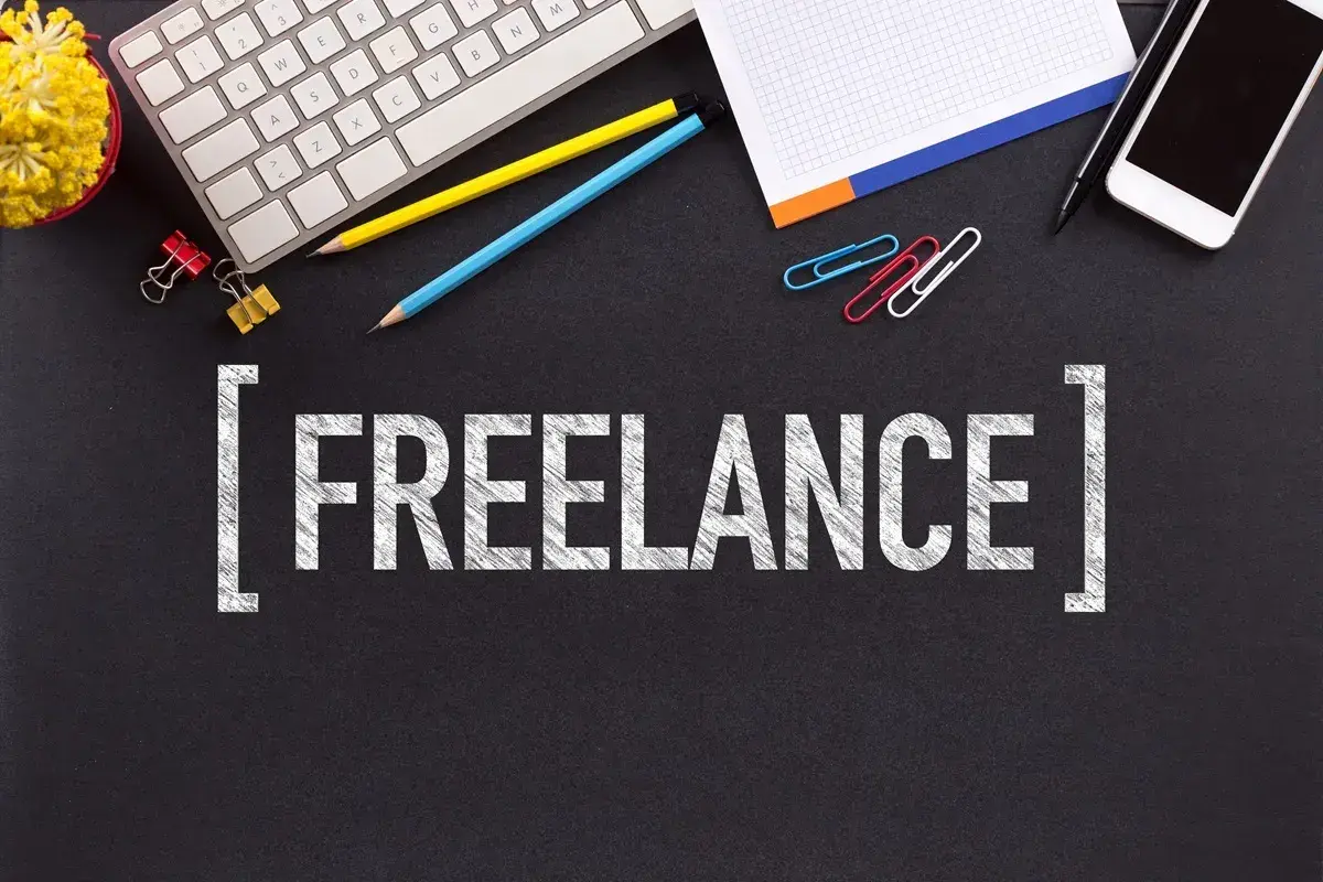 Biurko z klawiaturą, telefonem, notatnikiem i kredkami. Słowo "FREELANCE" na czarnym tle. To jest właśnie freelancer co to.