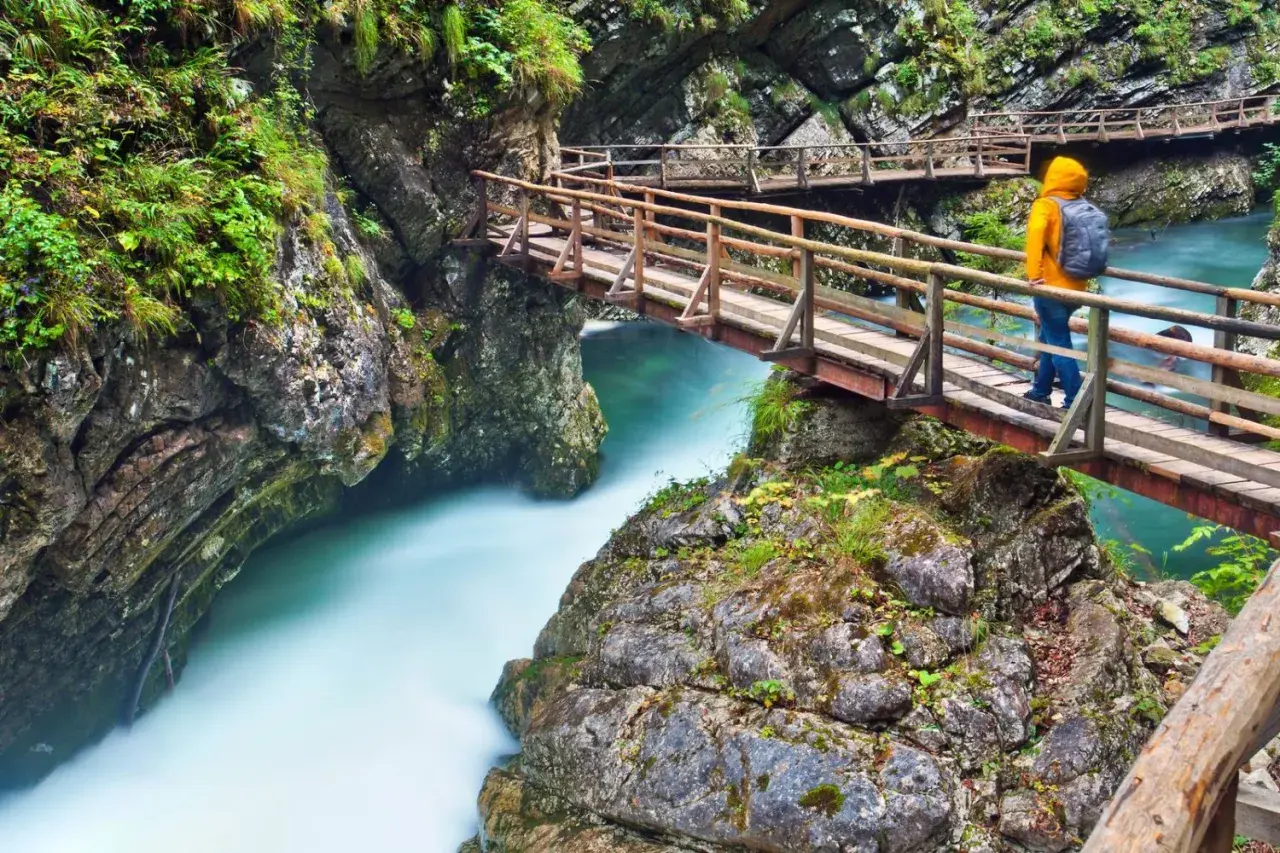 Ein Wanderer auf einer Holzbrücke über einem türkisfarbenen Fluss in einer Schlucht, ähnlich dem Bleder See.