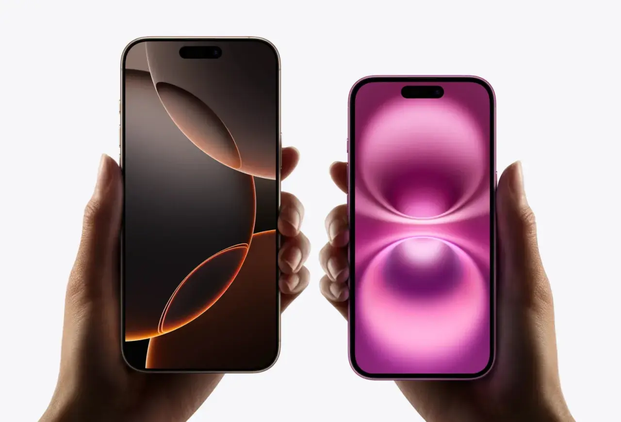 Dwie dłonie trzymają dwa iPhone'y. Jeden ma ciemny ekran z pomarańczowymi akcentami, drugi różowy. Oba modele mają dynamiczną wyspę.