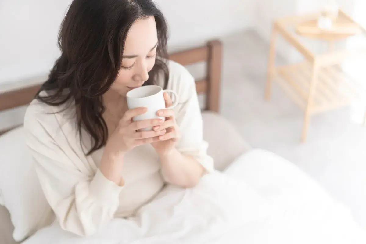 Donna con capelli scuri beve acqua calda al mattino, seduta a letto, avvolta in una coperta bianca.
