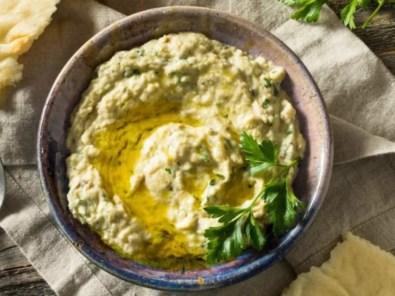 Baba ganush cremoso en un cuenco, con aceite de oliva y perejil fresco. Pan de pita al lado.