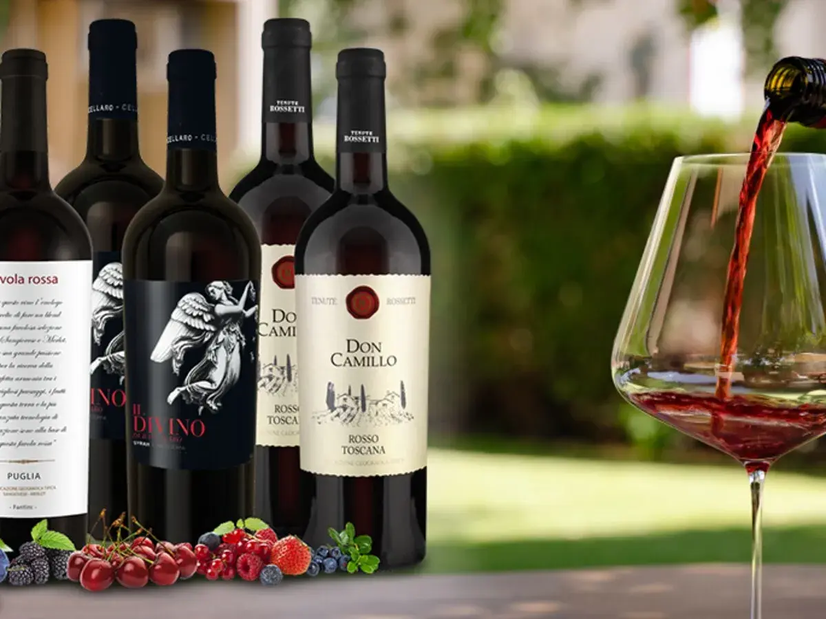 Flaschen mit teurem italienischem Rotwein, darunter "Il Divino" und "Don Camillo", werden vor einem Glas Rotwein präsentiert, in das gerade eingeschenkt wird. Frische Beeren und Kirschen liegen im Vordergrund.