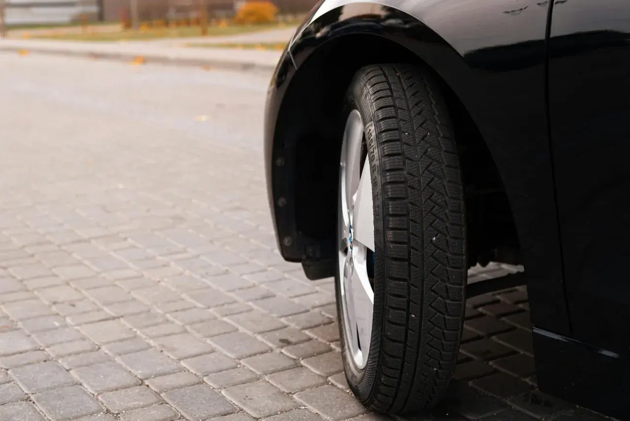 Błyszczące, czarne auto na kostce brukowej. Widoczna opona zimowa, której wiek można ocenić po bieżniku.