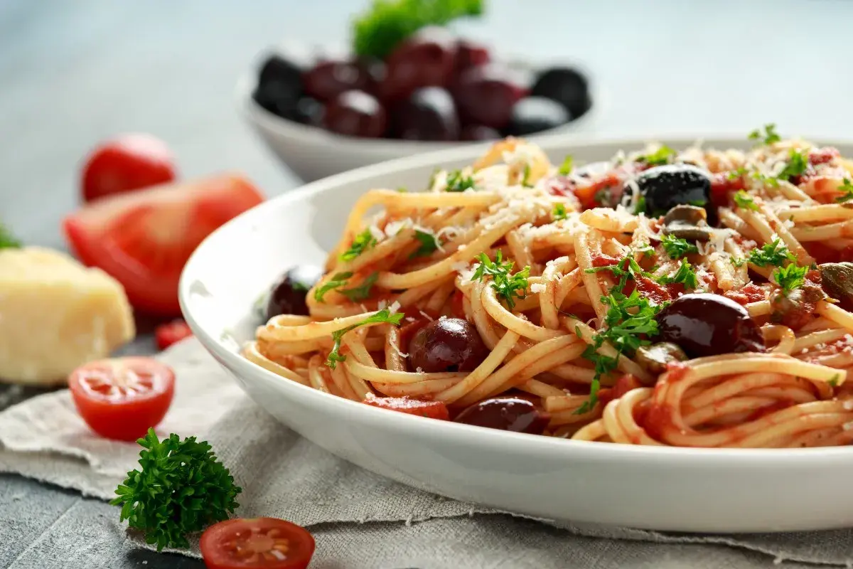 Un piatto di spaghetti alla puttanesca, conditi con pomodoro, olive e capperi, guarnito con prezzemolo e parmigiano.