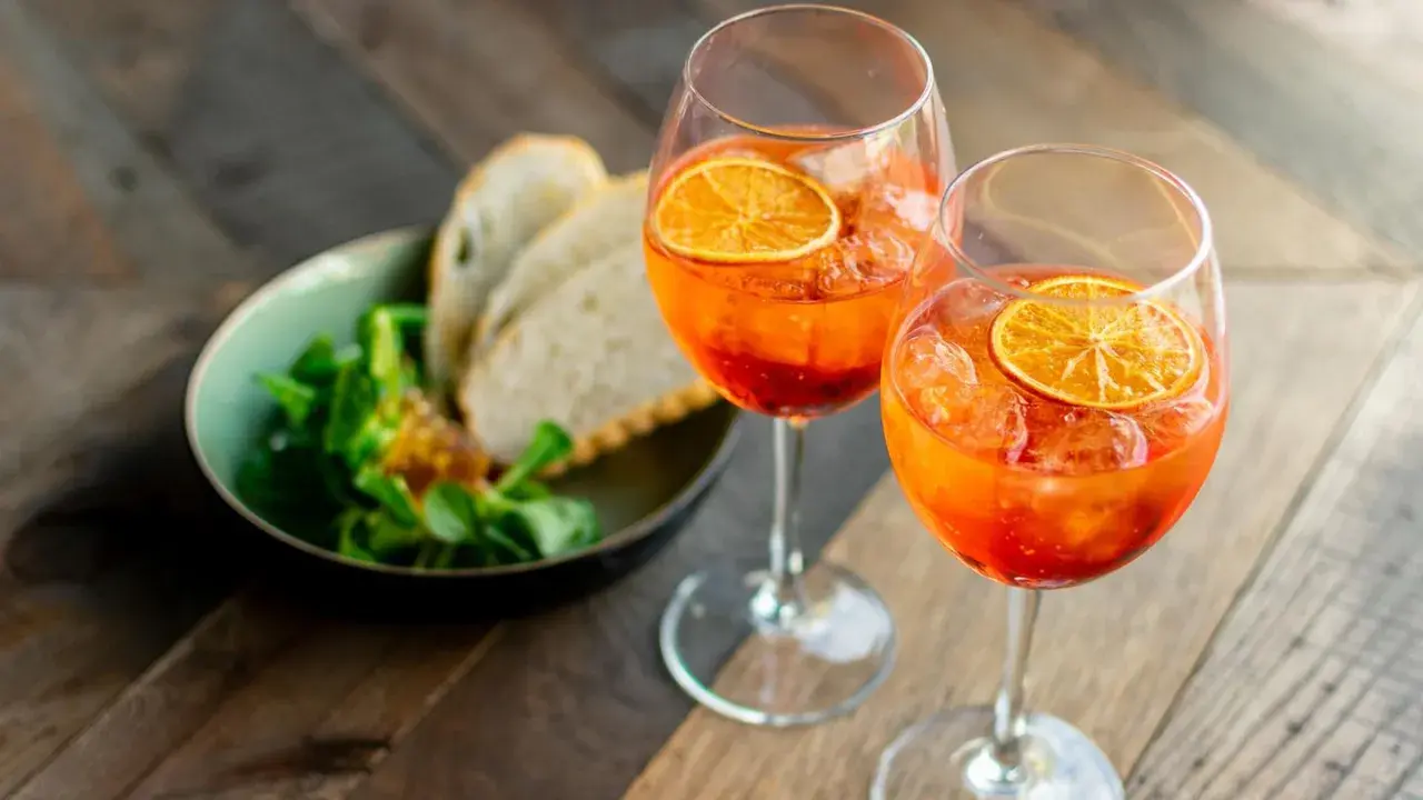 Zwei Gläser mit einem orangen Aperitif, garniert mit Orangenscheiben, daneben ein Schälchen mit Salat und Brot.
