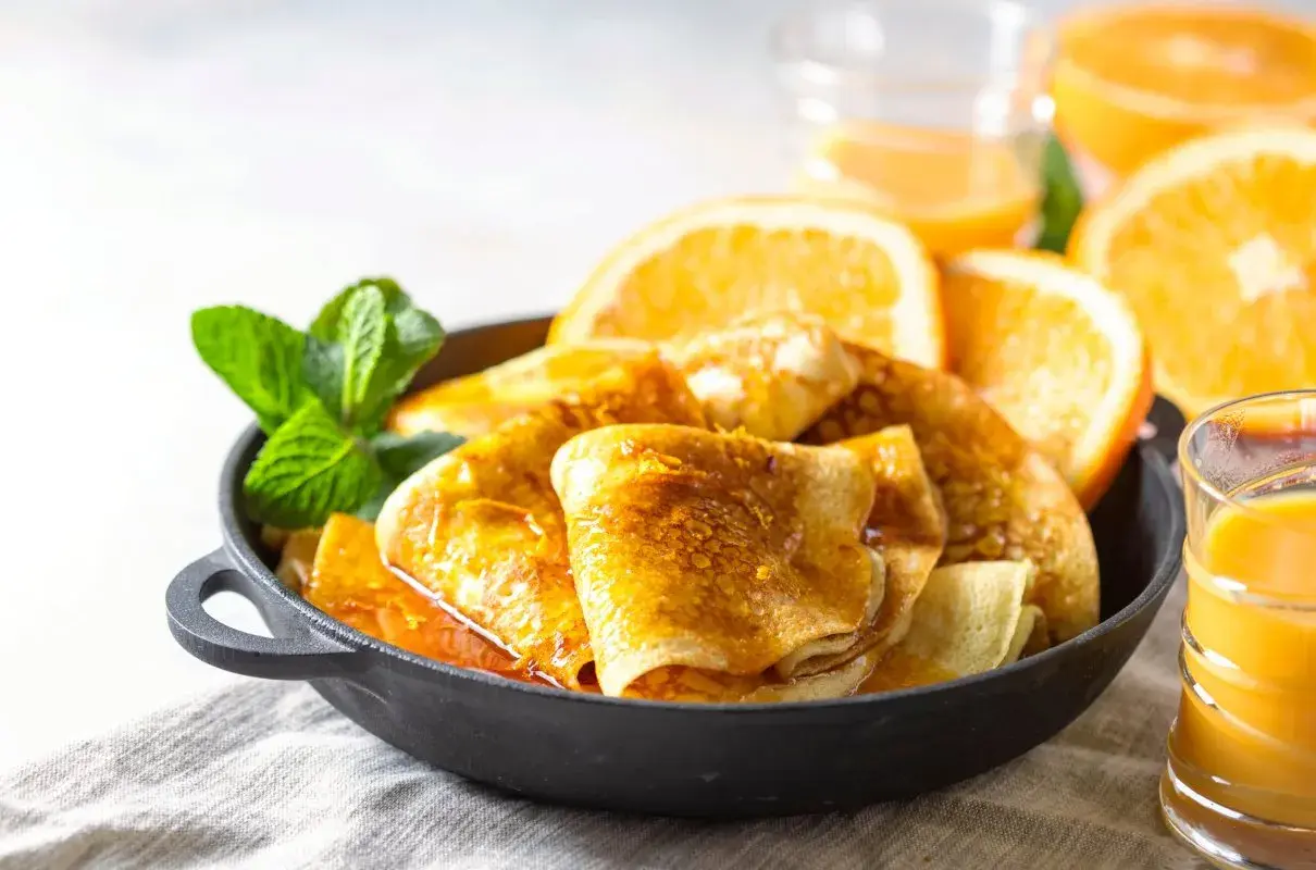Złociste crepes suzette w sosie pomarańczowym, udekorowane miętą i plasterkami pomarańczy, obok szklanki soku.