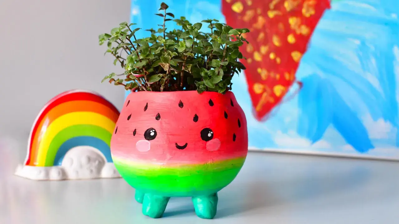Urocza doniczka diy w kształcie arbuza z uśmiechniętą buźką, pełna zielonych roślinek. Obok tęczowa ozdoba.