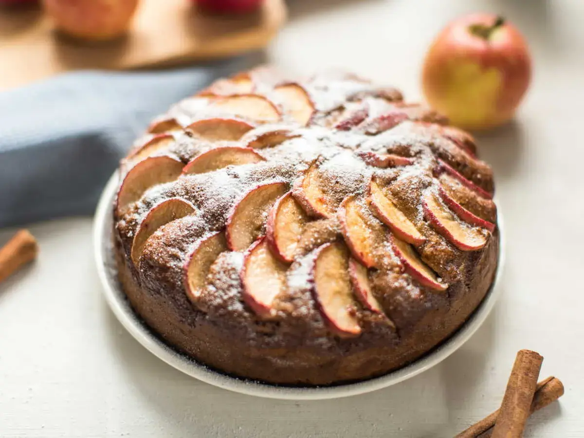 Un délicieux moelleux aux pommes, saupoudré de sucre glace, avec des pommes fraîches et des bâtons de cannelle en arrière-plan.