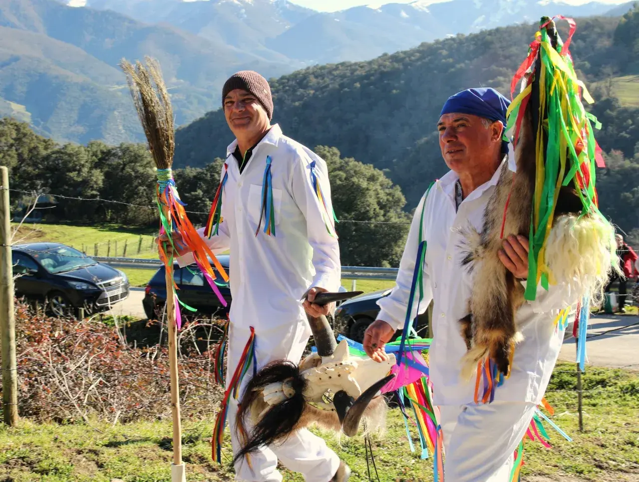 Hombres con trajes blancos y cintas de colores, portando una escoba y una cabeza de cabra, celebran tradiciones de Cantabria.