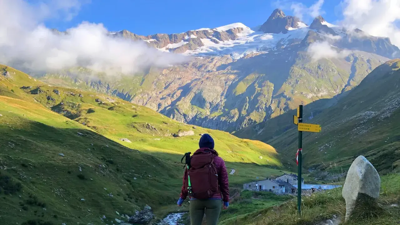 A hiker pauses on a **Tour Mont Blanc hike**, gazing at a majestic snow-capped mountain range. A signpost points the way to the next stage of this epic journey.