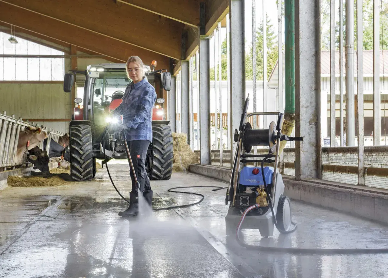 Frau reinigt Stallboden mit einem Industrie Hochdruckreiniger. Kühe und Traktor im Hintergrund.