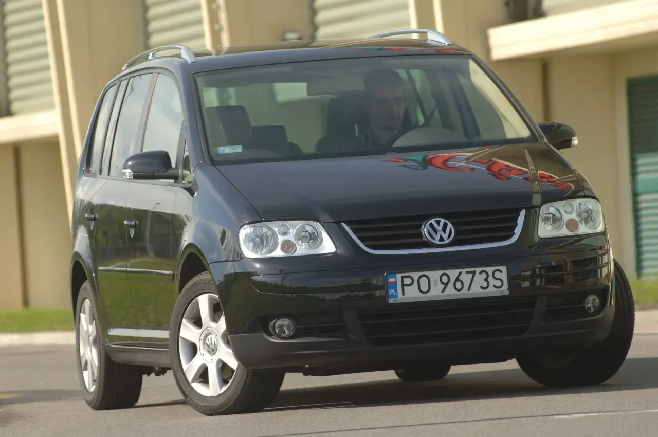 Czarny VW Touran z przodu, widoczne reflektory i żarówki. Kierowca za kierownicą.