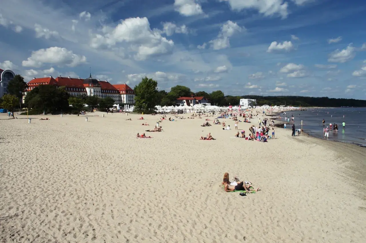 Plaża w Sopocie z ludźmi opalającymi się i spacerującymi, w tle budynki i morze.