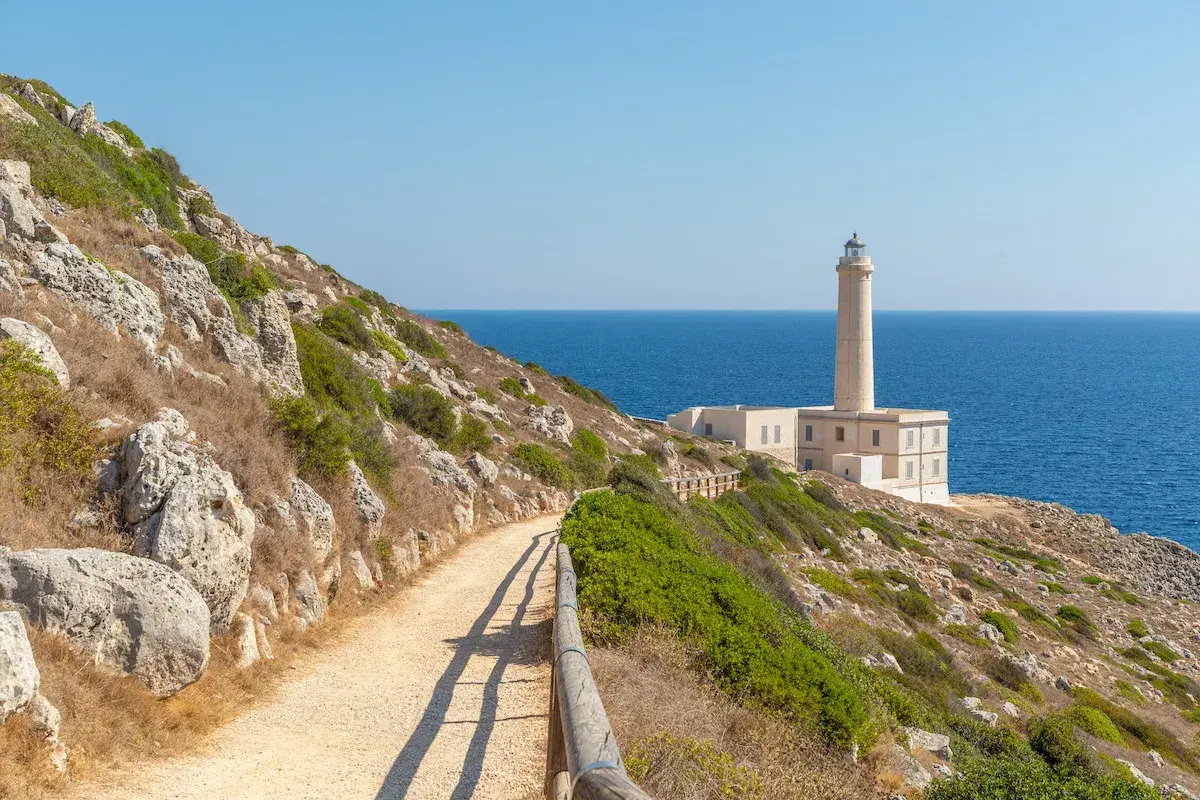 Sentiero roccioso con vegetazione mediterranea che porta a un faro bianco sul mare blu, tipico paesaggio del Salento città.