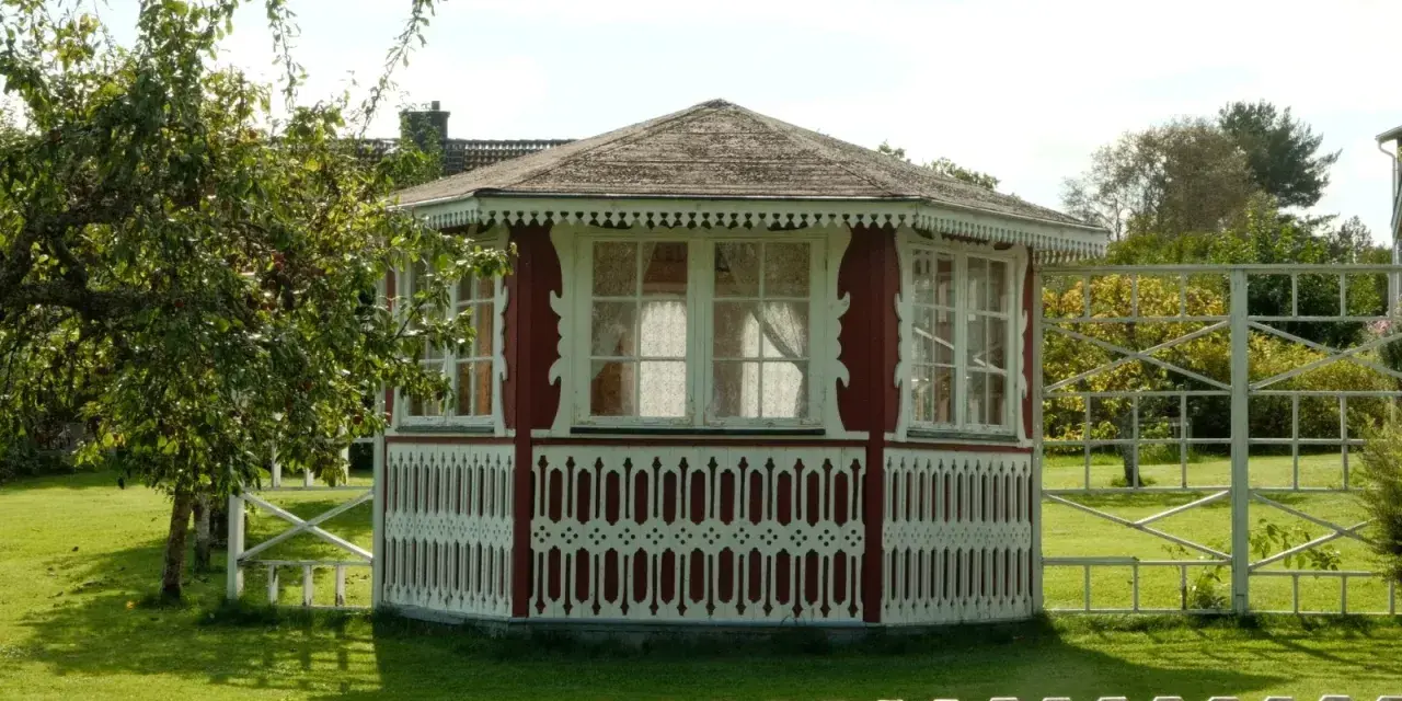 Pavillon de jardin rouge et blanc, orné de dentelle de bois. Une pomme tombe d'un arbre, peut-être pour échapper à la taxe abri de jardin.