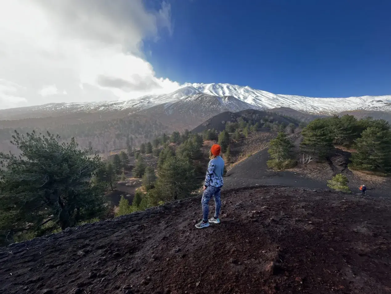 Kobieta w pomarańczowej czapce patrzy na ośnieżony wulkan Etna, otoczony lasem i czarną ziemią.