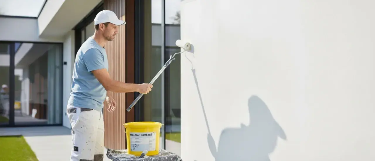 Mann streicht mit einer Fassadenrolle eine weiße Wand. Ein Eimer mit Farbe steht daneben.