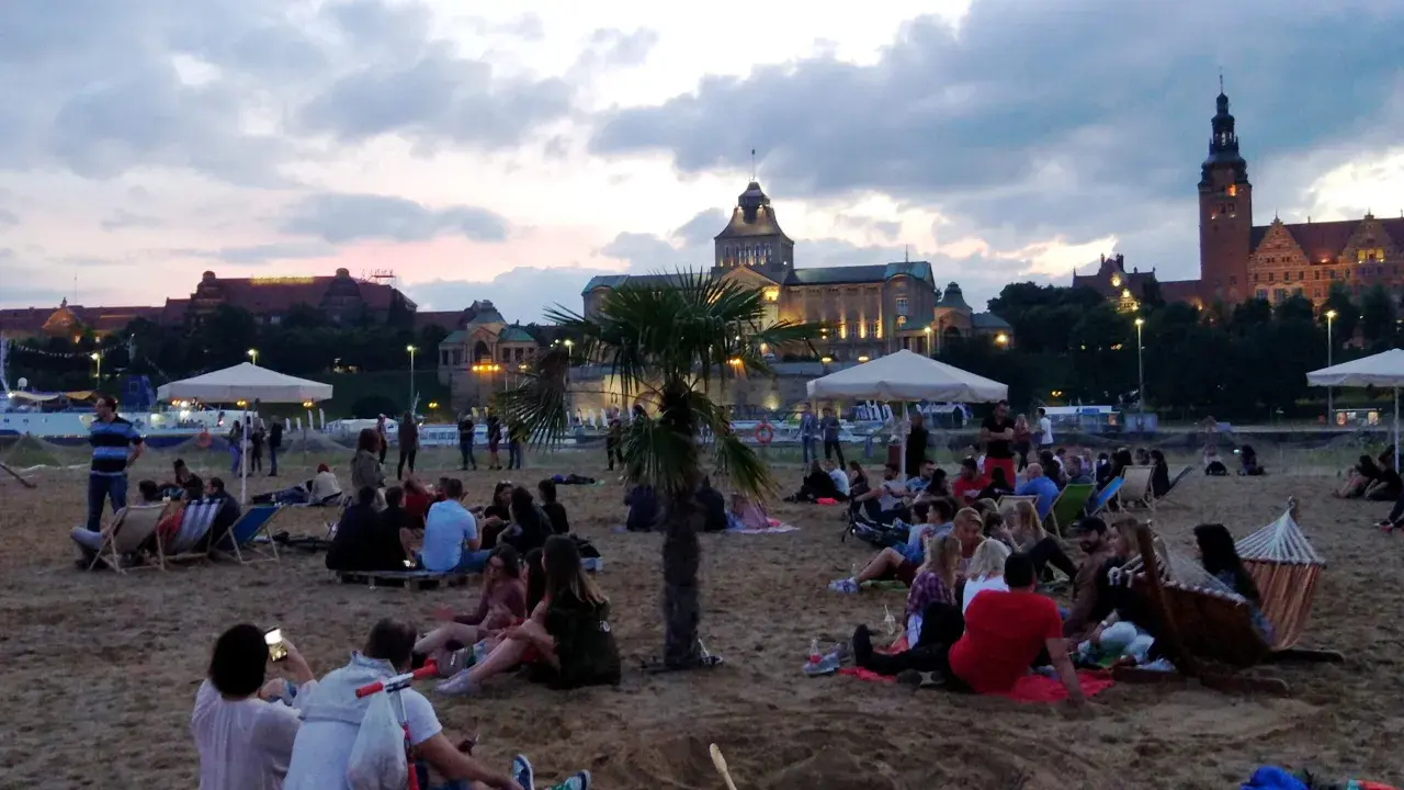 Ludzie odpoczywają na plaży, ciesząc się wieczorem. W tle widać piękne budynki, świadczące o bogactwie wydarzeń kulturalnych w Szczecinie.