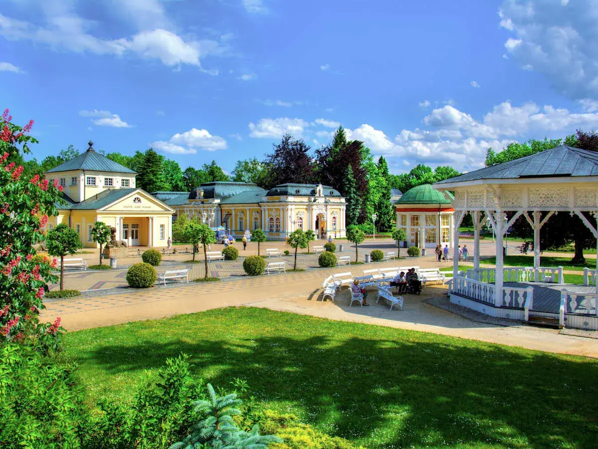 Ein sonniger Tag in einer **Therme Tschechien**. Gelbe Kurgebäude mit grünen Dächern, ein weißer Pavillon und gepflegte Gärten laden zum Entspannen ein.