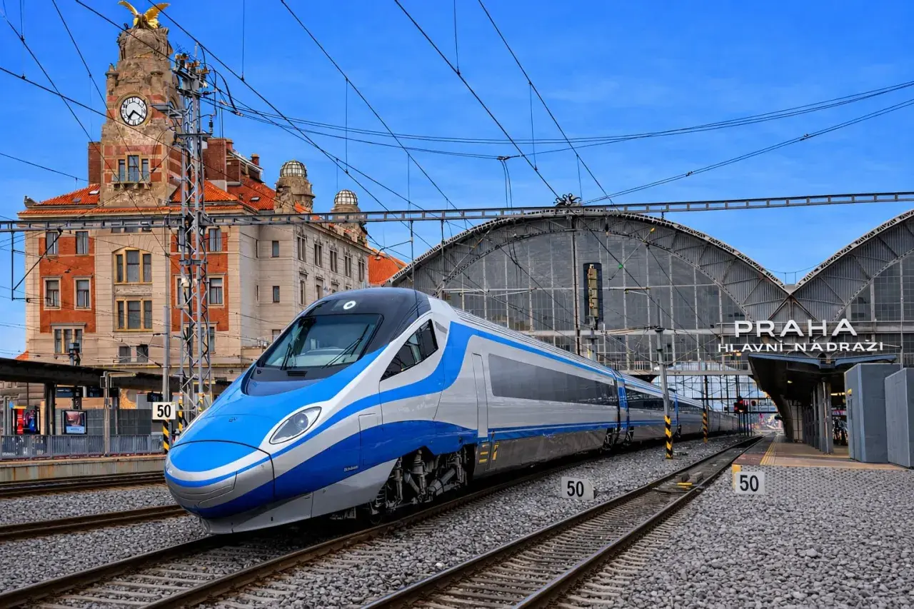 Nowoczesne oznaczenie Pendolino na tle zabytkowego dworca w Pradze. Pociąg w kolorach niebieskim i srebrnym stoi na torach.