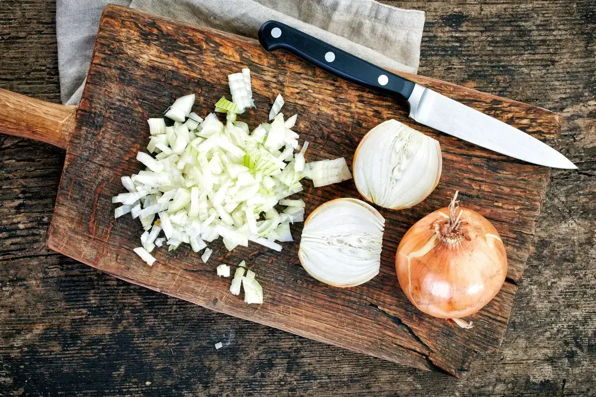 Cipolla cruda digeribile, tagliata finemente su un tagliere di legno, accanto a una cipolla intera e a metà. Un coltello è pronto all'uso.