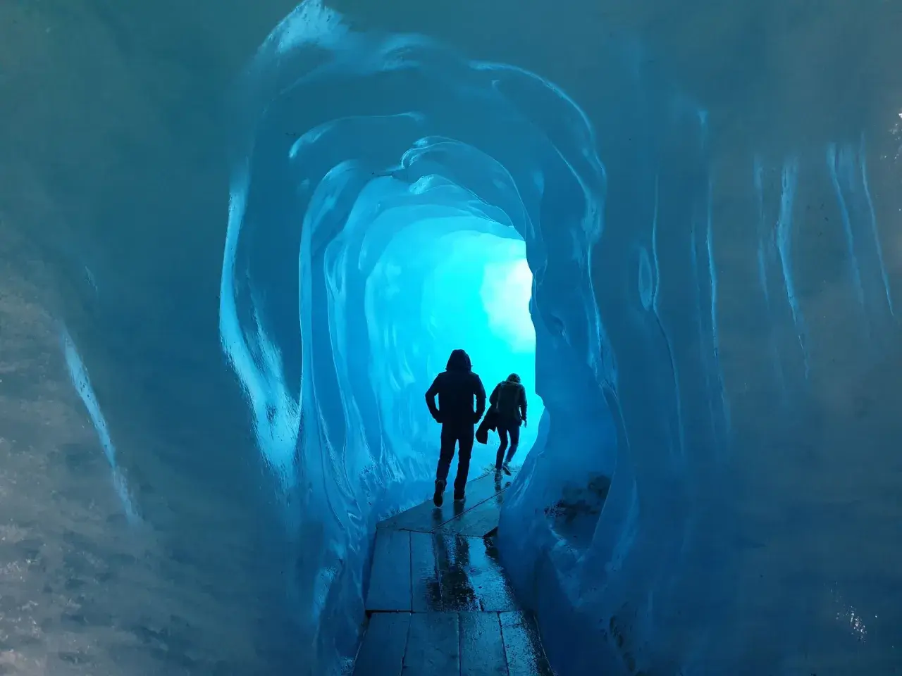 Dwie sylwetki wędrują przez lodowy tunel. Jasne, turkusowe światło przebija się z głębi, tworząc magiczną atmosferę. To jak podróż do wnętrza lodowca, gdzie czas się zatrzymał, a jedynym dźwiękiem jest echo kroków.