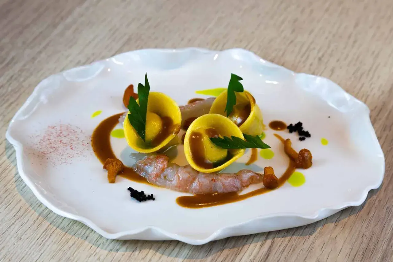 Trois raviolis italiens dorés, garniture de crevettes, champignons et sauce brune sur une assiette blanche ondulée.