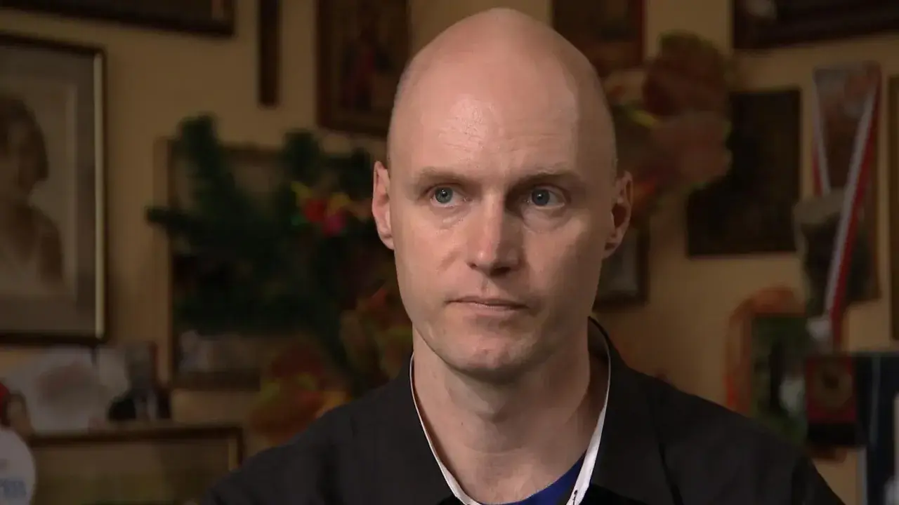 Krzysztof Igor Krawczyk, łysy mężczyzna z niebieskimi oczami, w czarnej koszuli, na tle rodzinnych zdjęć.