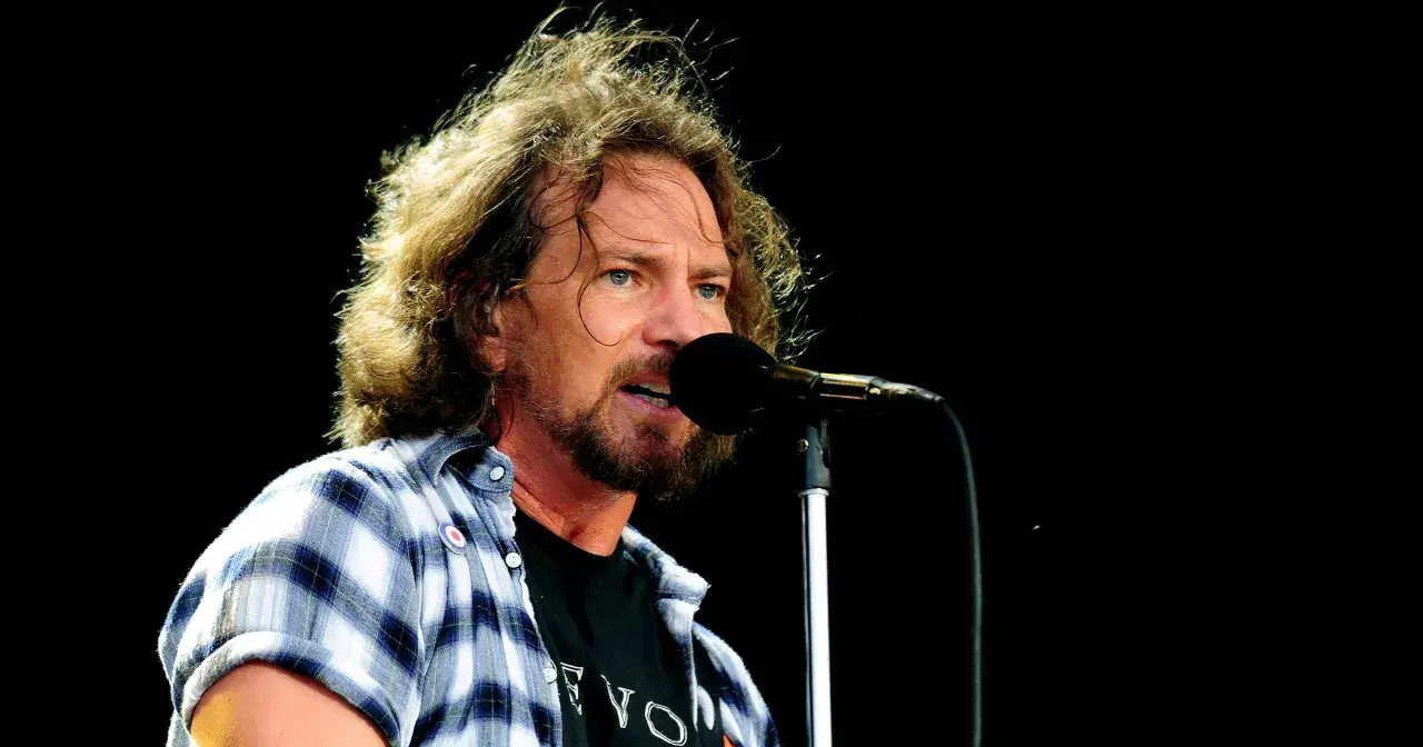 Eddie Vedder, wokalista Pearl Jam, śpiewa do mikrofonu na koncercie. Jego rozwichrzone włosy i charakterystyczna koszula dodają mu rockowego stylu.