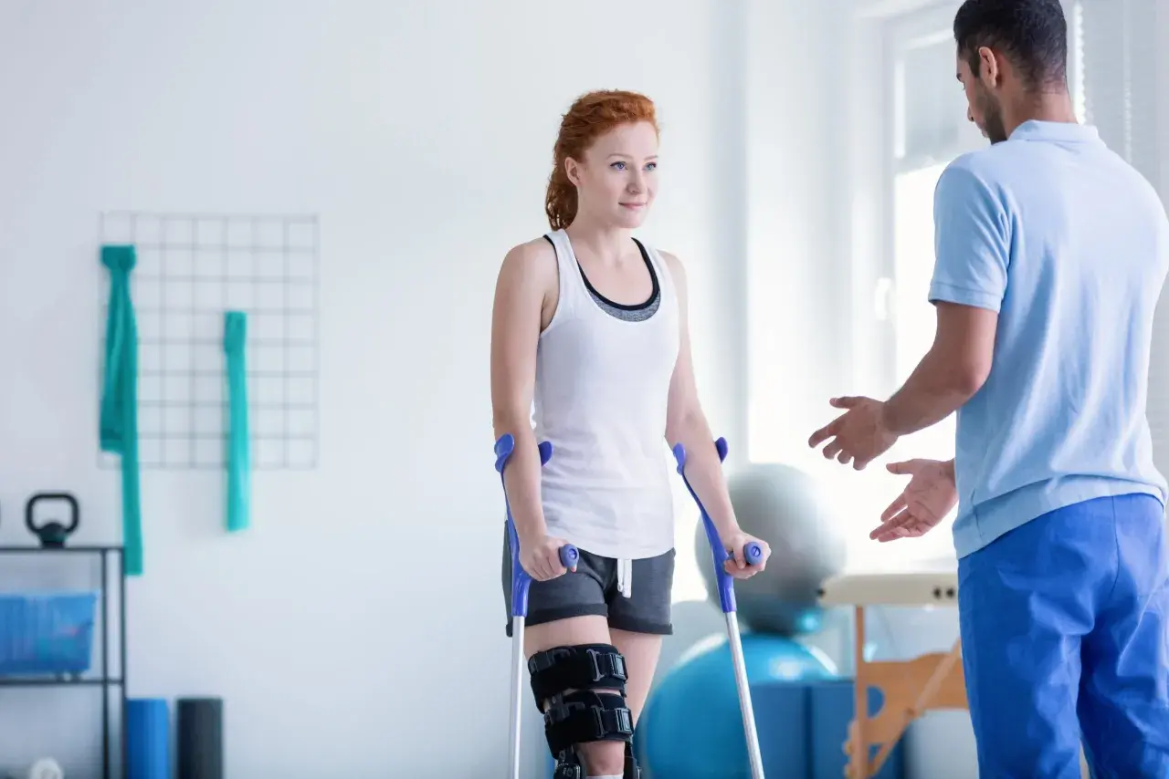 Kobieta na kulach z ortezą na nodze podczas zabiegów rehabilitacyjnych, wspierana przez fizjoterapeutę.