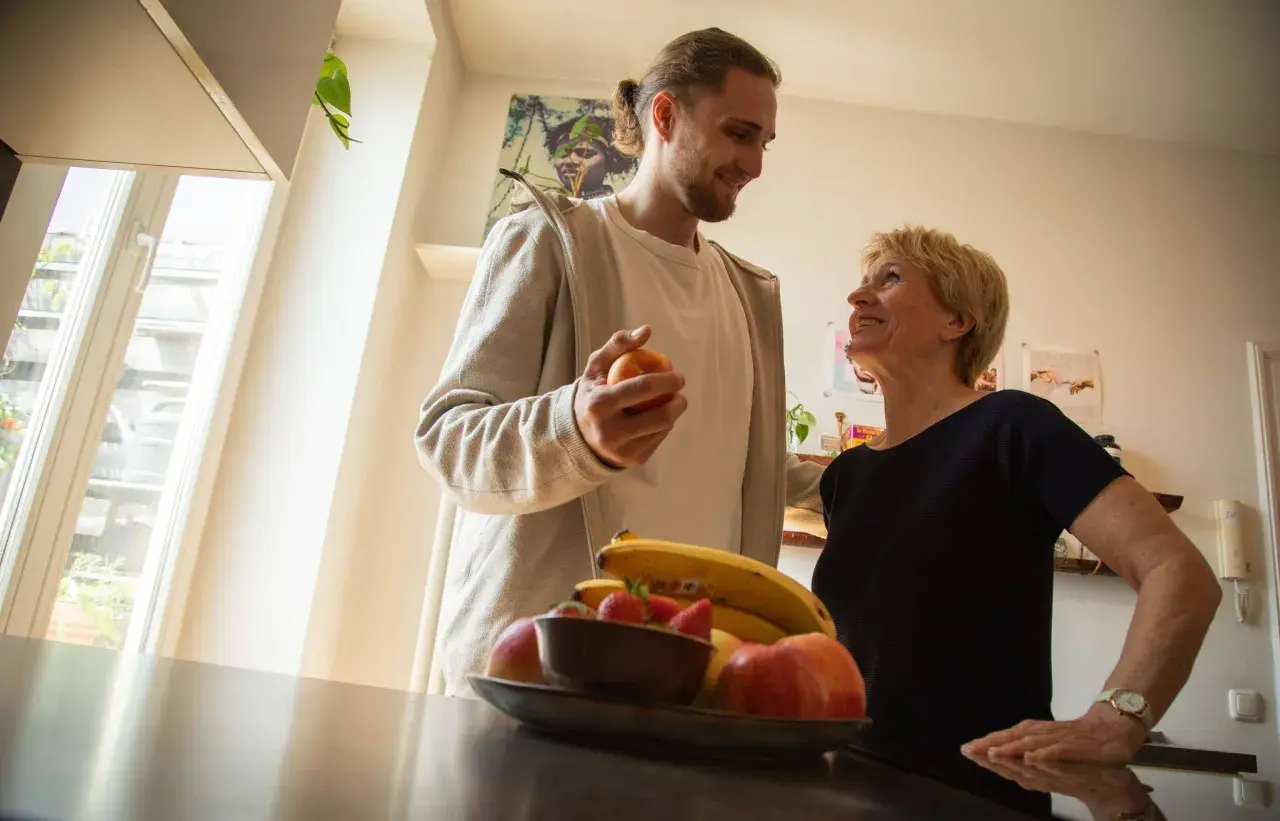Ein Mann hält einen Apfel, während er mit einer lächelnden Frau spricht. Obst liegt auf einem Tisch. Vielleicht ein Moment der Entlastung für eine Privatperson.