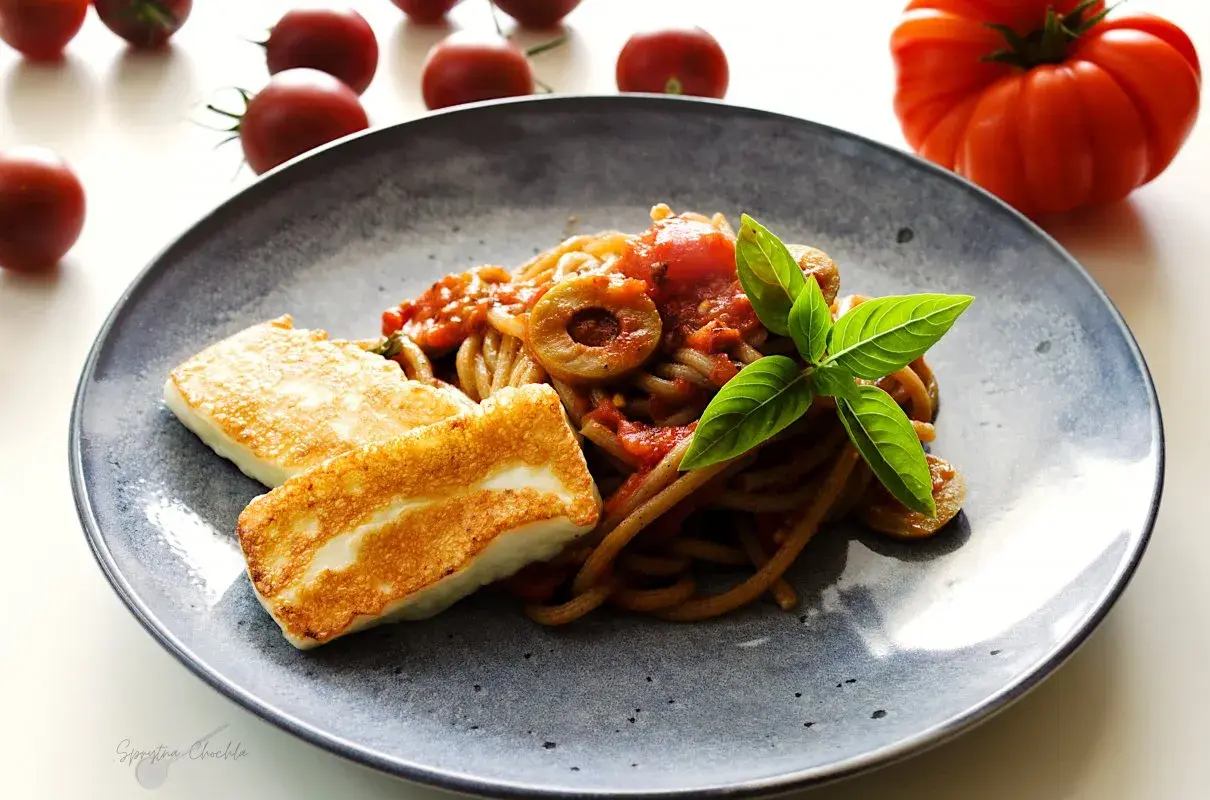 Makaron z halloumi na talerzu, z sosem pomidorowym, oliwkami i bazylią. Obok leżą świeże pomidory.
