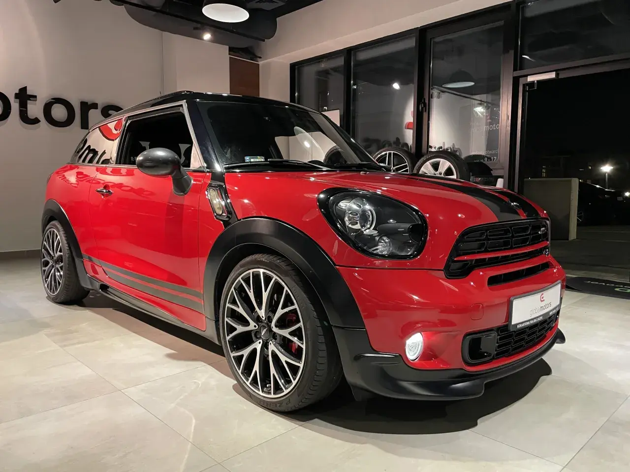 Czerwony mini paceman z czarnymi pasami i felgami stoi w salonie.