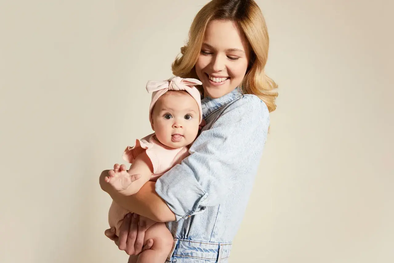 Uśmiechnięta mama tuli niemowlę w różowym body i opasce. Wskazówka, jak ubierać noworodka jesienią, by było mu ciepło i wygodnie.