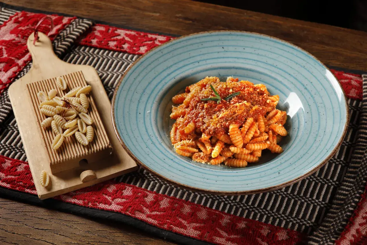 Gnocchetti sardi conditi con sugo rosso e parmigiano, accanto a una spianatoia con pasta fresca.