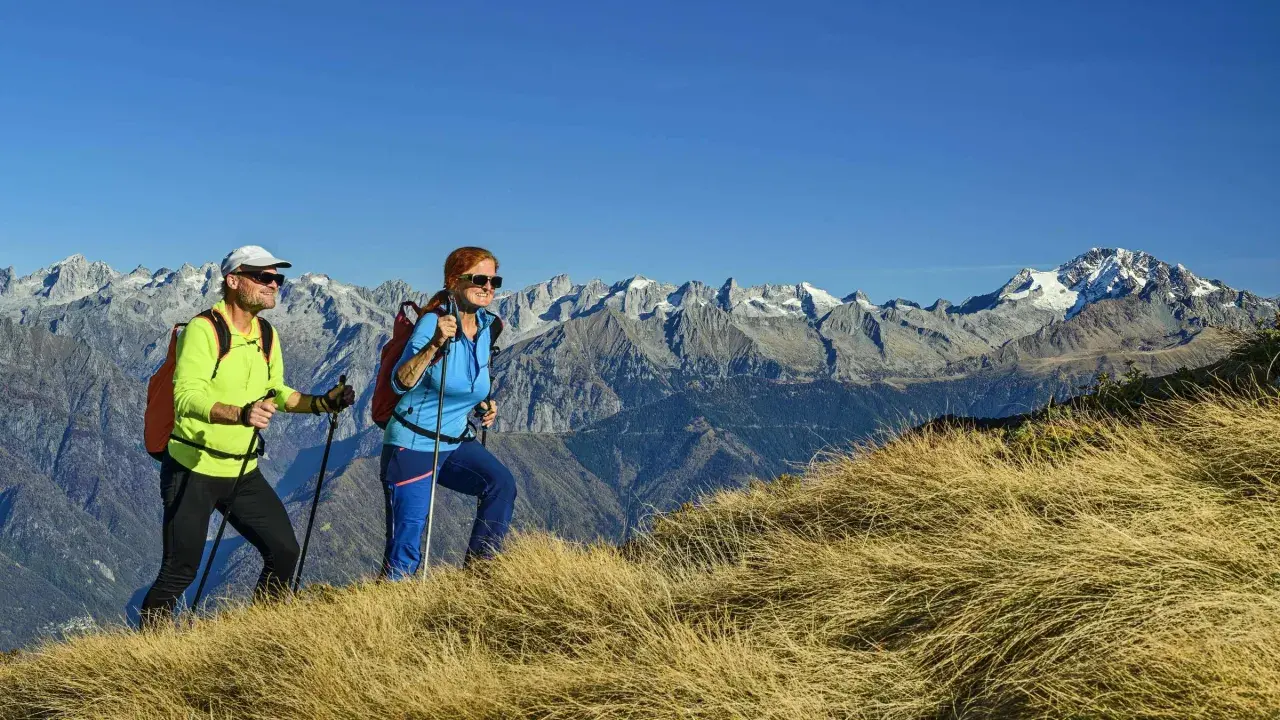 Zwei Wanderer genießen die Aussicht auf die Alpen. Sie gehen auf einem grasbewachsenen Hang, Stöcke in den Händen, mit schneebedeckten Gipfeln im Hintergrund.