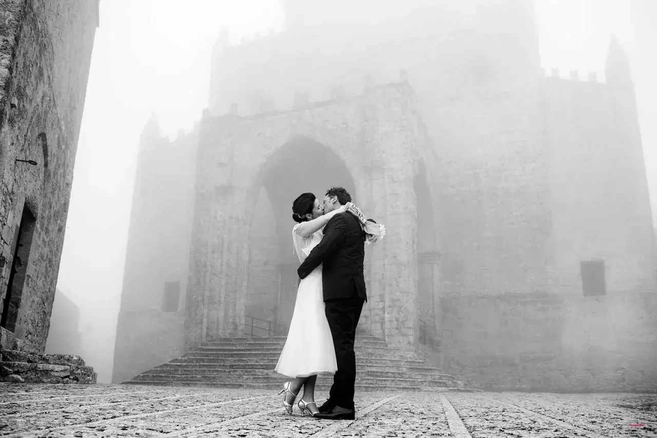 Sposi si baciano davanti a un antico arco in pietra, immersi nella nebbia. Dettagli matrimonio suggestivi.