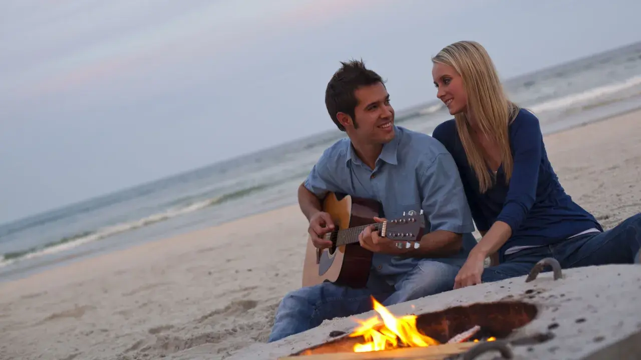 Coppia sulla spiaggia al tramonto, lui suona la chitarra, lei lo guarda sorridendo. Un falò illumina la scena, ma la noia nella coppia è palpabile.